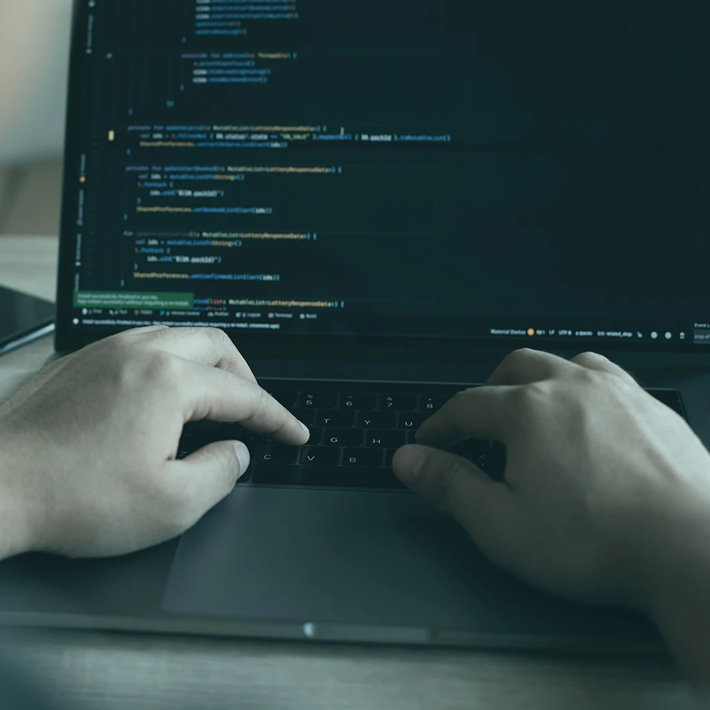 Hände tippen auf Laptop-Tastatur, Programmier-Umgebung ist am Laptop-Bildschirm zu sehen.